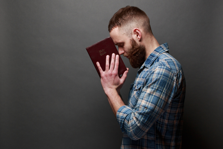 Handsome man reading and praying over Bible in a dark room over gray texture