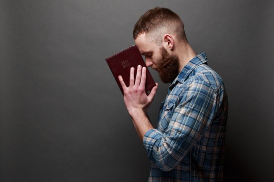 Handsome man reading and praying over Bible in a dark room over gray texture